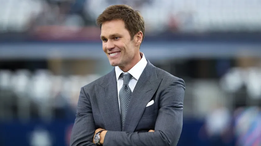 Tom Brady is pictured standing in a stadium, wearing a gray suit with a white dress shirt and a patterned tie. He is smiling with his arms crossed. The background is blurred, showing a large, open space with stadium seating.